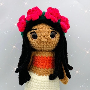 Puede incluir: Muñeca de crochet con piel marrón, cabello negro y falda blanca. La muñeca lleva un top rojo y una corona de flores verde y rosa.