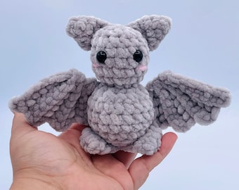 May include: A gray crocheted bat plush toy with black eyes and pink cheeks. The bat has large wings and is sitting on a hand.