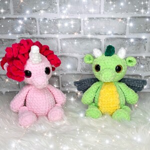 Puede incluir: Un unicornio de crochet rosa y un dragón de crochet verde con alas grises. Ambos están sentados sobre una superficie blanca y esponjosa.