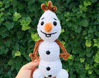 May include: A white crocheted snowman with brown yarn hair and an orange carrot nose. The snowman is smiling and has black eyes and buttons.