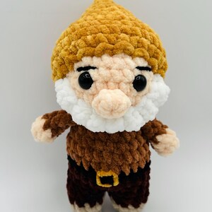 May include: A handmade crochet gnome doll with a yellow hat, white beard, and brown outfit. The gnome has black eyes and a small nose. The outfit includes a brown top and pants with a gold buckle. This whimsical doll is perfect for home decor.