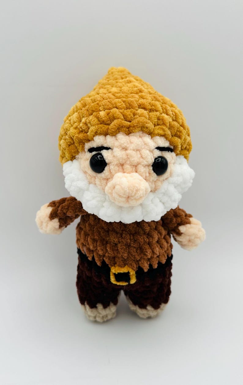 May include: A handmade crochet gnome doll with a yellow hat, white beard, and brown outfit. The gnome has black eyes and a small nose. The outfit includes a brown top and pants with a gold buckle. This whimsical doll is perfect for home decor.