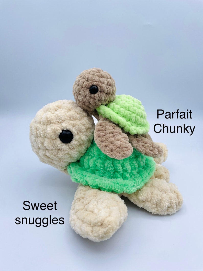 May include: Two crocheted plush turtle toys, one larger than the other, with green shells and brown bodies. The smaller turtle is riding on the back of the larger turtle. The larger turtle has the text "Sweet snuggles" written below it. The smaller turtle has the text "Parfait Chunky" written above it.