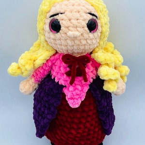 K&ouml;nnte beinhalten: Eine geh&auml;kelte Puppe mit langen gelben Haaren, die ein rosa-violettes Kleid mit einer roten Schleife tr&auml;gt. Die Puppe hat gro&szlig;e schwarze Augen mit rosa Glitzer.