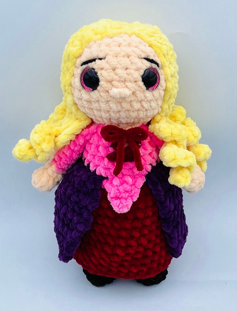 K&ouml;nnte beinhalten: Eine geh&auml;kelte Puppe mit langen gelben Haaren, die ein rosa-violettes Kleid mit einer roten Schleife tr&auml;gt. Die Puppe hat gro&szlig;e schwarze Augen mit rosa Glitzer.