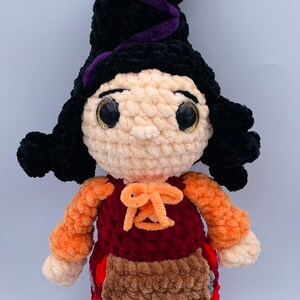 K&ouml;nnte beinhalten: Eine geh&auml;kelte Hexenpuppe mit schwarzen Haaren, einem spitzen Hut in Lila und Schwarz, einem roten und orangenen Kleid und einer braunen Sch&uuml;rze.