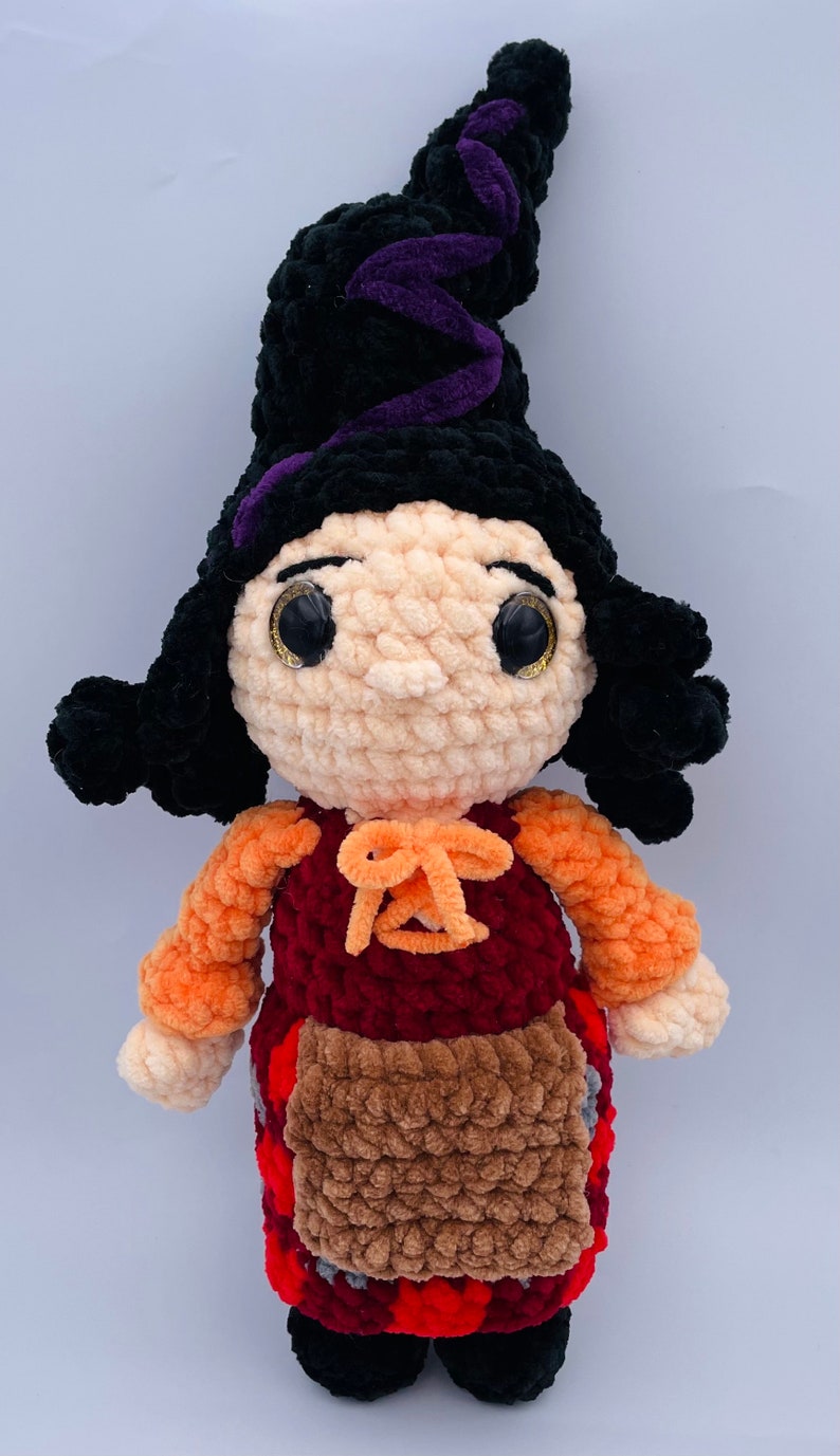 K&ouml;nnte beinhalten: Eine geh&auml;kelte Hexenpuppe mit schwarzen Haaren, einem spitzen Hut in Lila und Schwarz, einem roten und orangenen Kleid und einer braunen Sch&uuml;rze.