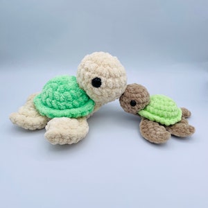 May include: Two crocheted plush turtle toys. The larger turtle is cream colored with a green shell. The smaller turtle is brown with a green shell. The turtles are facing each other and appear to be hugging.