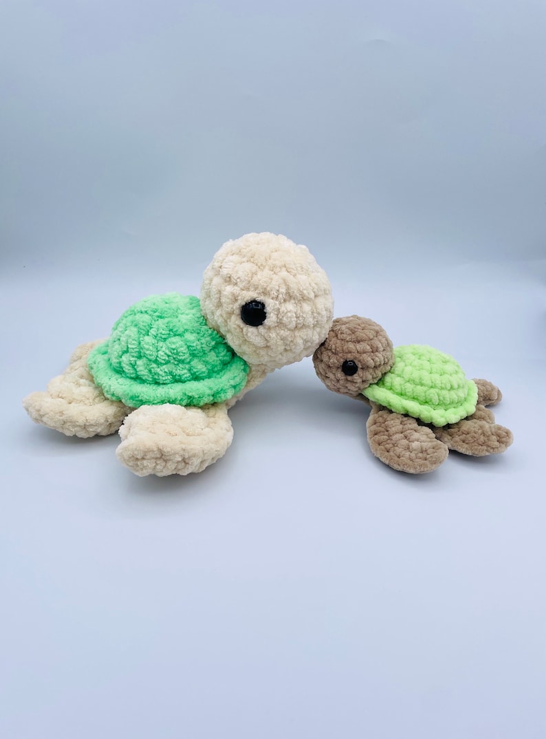 May include: Two crocheted plush turtle toys. The larger turtle is cream colored with a green shell. The smaller turtle is brown with a green shell. The turtles are facing each other and appear to be hugging.