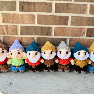 Puede incluir: Siete muñecos de gnomos hechos a mano con ganchillo, con sombreros, camisas y cinturones de varios colores. Cada gnomo tiene una barba blanca y una cara sonriente. Los muñecos están colocados en fila contra una pared de ladrillos.