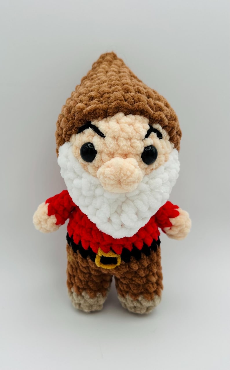 May include: A handmade crochet dwarf doll with a brown hat, face, and legs. The doll has a white beard, red shirt, and black belt with a gold buckle. The eyes are large and black.