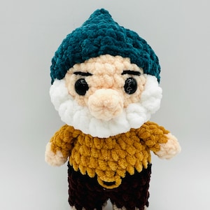 May include: A handmade crochet gnome doll with a teal hat, a white beard, and a tan face. The gnome wears a mustard yellow top and brown pants. The doll has black button eyes and is made of soft, textured yarn.
