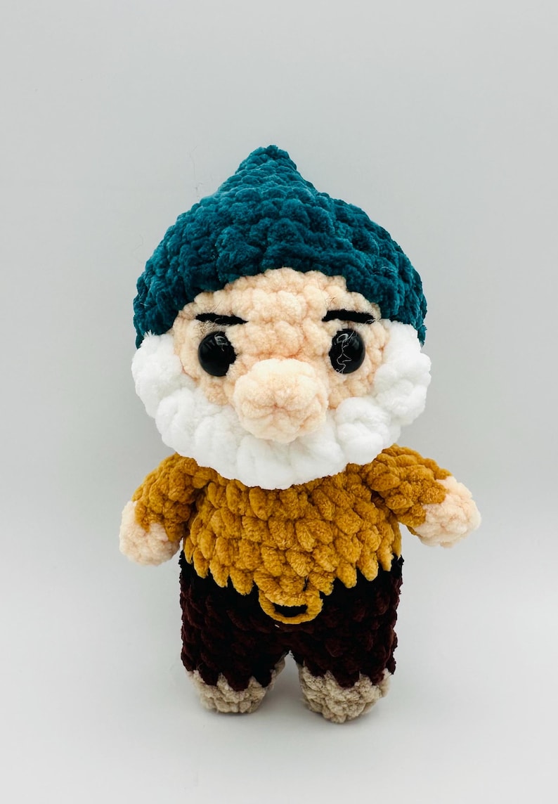 May include: A handmade crochet gnome doll with a teal hat, a white beard, and a tan face. The gnome wears a mustard yellow top and brown pants. The doll has black button eyes and is made of soft, textured yarn.