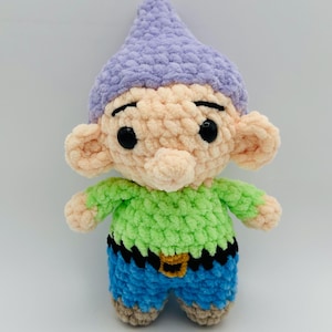 May include: A handmade crochet doll of a cartoon character. The doll has a light purple hat, peach-colored face and ears, a green shirt, blue shorts, and black eyes. The doll has a black belt with a gold buckle.