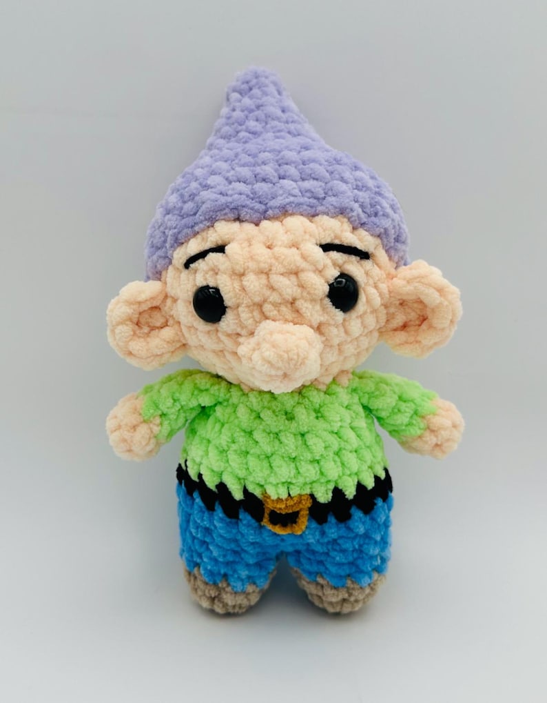 May include: A handmade crochet doll of a cartoon character. The doll has a light purple hat, peach-colored face and ears, a green shirt, blue shorts, and black eyes. The doll has a black belt with a gold buckle.