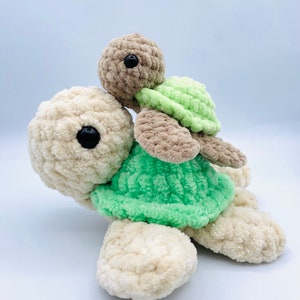 May include: Two crocheted plush turtles, one riding on top of the other. The larger turtle is beige with a green shell, and the smaller turtle is brown with a green shell.
