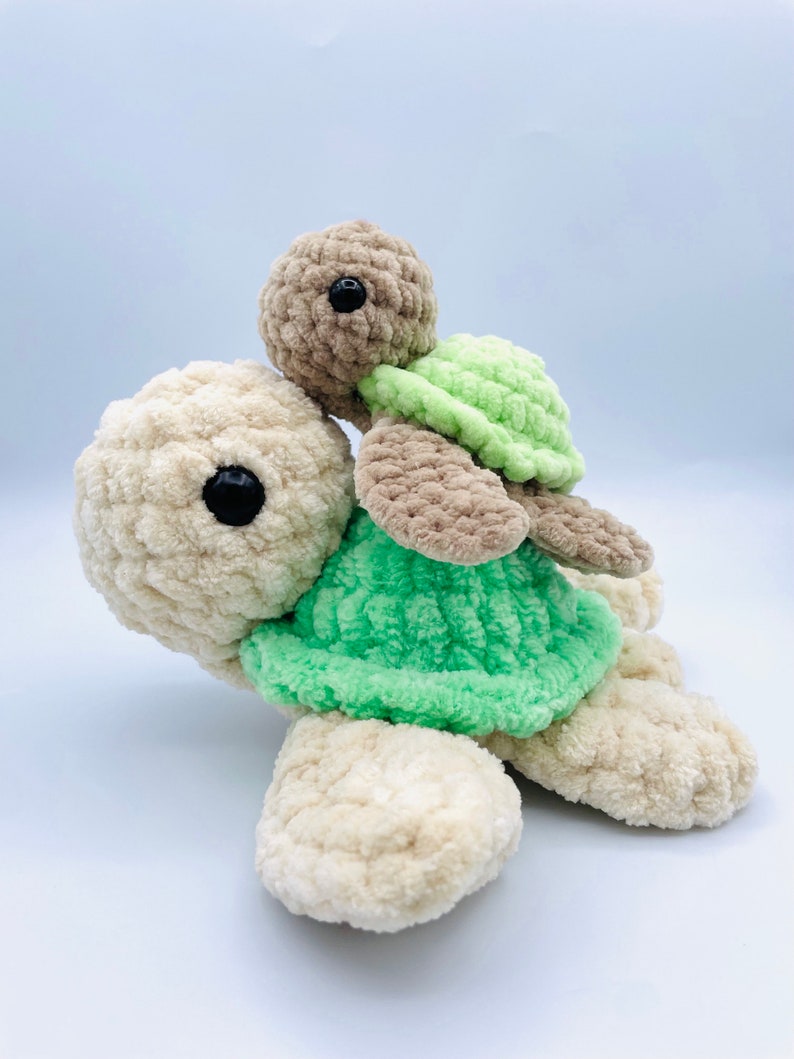 May include: Two crocheted plush turtles, one riding on top of the other. The larger turtle is beige with a green shell, and the smaller turtle is brown with a green shell.