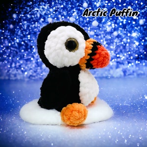 Puede incluir: Un juguete de peluche de frailecillo ártico de crochet con pelaje blanco y negro, pico y patas naranjas y grandes ojos negros. El frailecillo está sentado sobre una superficie blanca de crochet.