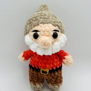 May include: A handmade crochet gnome doll with a tan hat, peach face, and white beard. The gnome wears a red shirt, brown pants, and a black belt with a gold buckle. The doll is a whimsical decorative item.