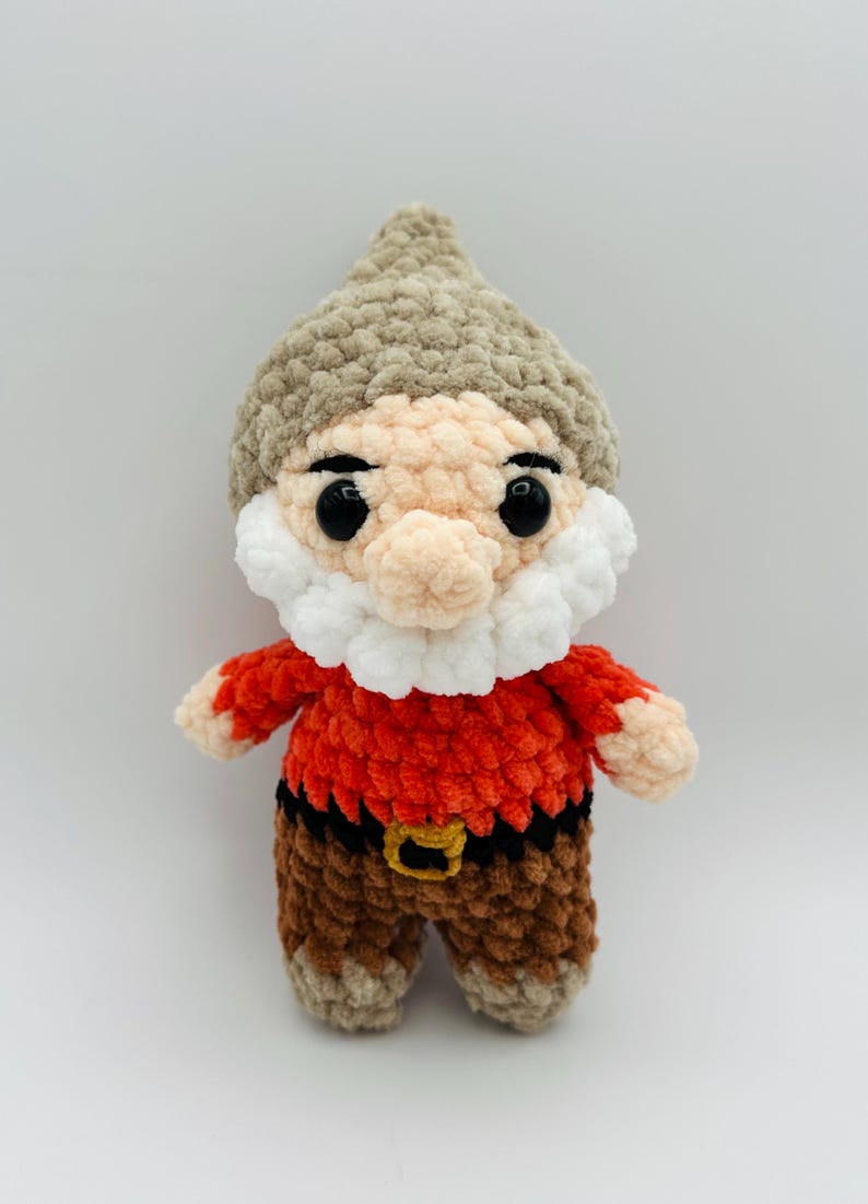 May include: A handmade crochet gnome doll with a tan hat, peach face, and white beard. The gnome wears a red shirt, brown pants, and a black belt with a gold buckle. The doll is a whimsical decorative item.