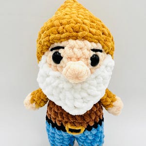 May include: A handmade crochet gnome doll with a yellow hat, white beard, and blue pants. The gnome has a brown top and a black belt with a gold buckle. The doll has black eyes and a pink nose.