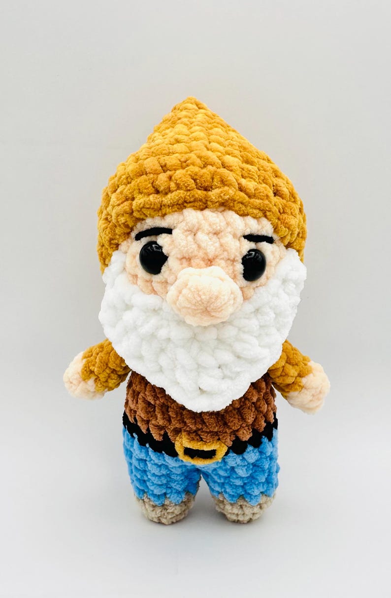 May include: A handmade crochet gnome doll with a yellow hat, white beard, and blue pants. The gnome has a brown top and a black belt with a gold buckle. The doll has black eyes and a pink nose.
