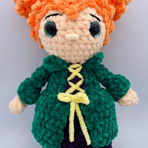 K&ouml;nnte beinhalten: Eine geh&auml;kelte Puppe mit orangefarbenem Haar, gr&uuml;ner Jacke, lila Kleid und gelbem Band. Die Puppe hat gro&szlig;e gr&uuml;ne Augen und einen s&uuml;&szlig;en Ausdruck.