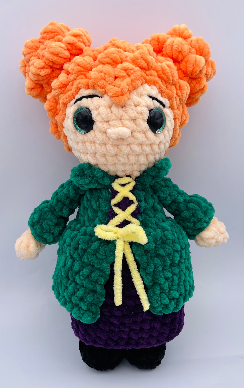 K&ouml;nnte beinhalten: Eine geh&auml;kelte Puppe mit orangefarbenem Haar, gr&uuml;ner Jacke, lila Kleid und gelbem Band. Die Puppe hat gro&szlig;e gr&uuml;ne Augen und einen s&uuml;&szlig;en Ausdruck.