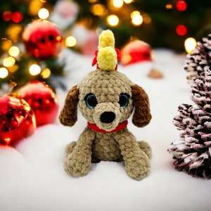 Puede incluir: Un juguete de perro de crochet con pelaje marrón, ojos azules y un collar rojo. El juguete lleva una diadema de reno de crochet amarilla y marrón. El juguete está sentado sobre una superficie blanca con un fondo borroso de luces navideñas y adornos.