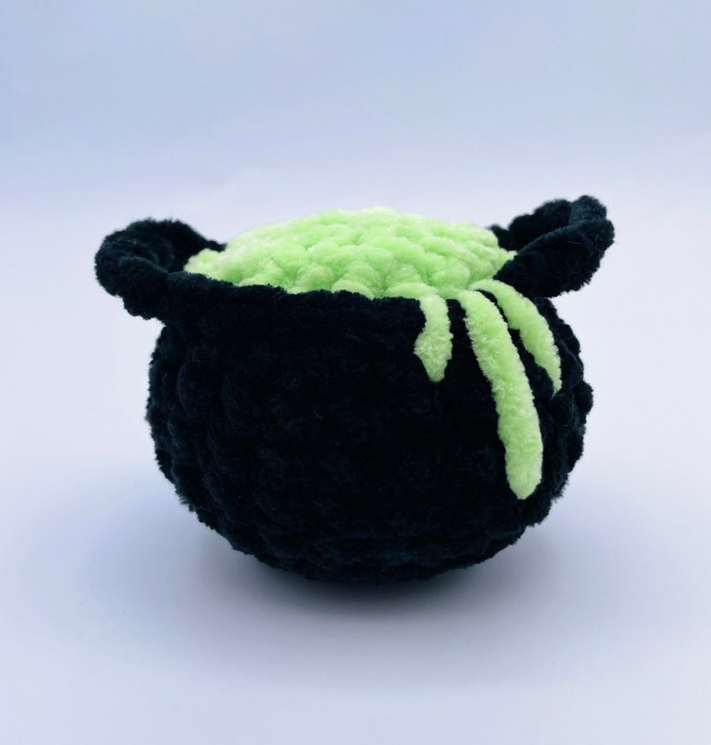 May include: A black crocheted cauldron filled with green slime. The slime is dripping down the sides of the cauldron.