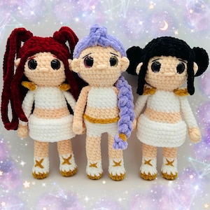 May include: Three handmade crochet dolls with red, purple, and black hair. Each doll has a cream-coloured face, white dress, and gold accents. The dolls are standing in front of a starry background.