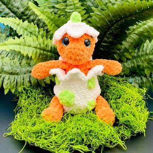 May include: A crocheted orange dinosaur toy with green spots, emerging from a white egg with green spots. The dinosaur is sitting on a bed of green moss.