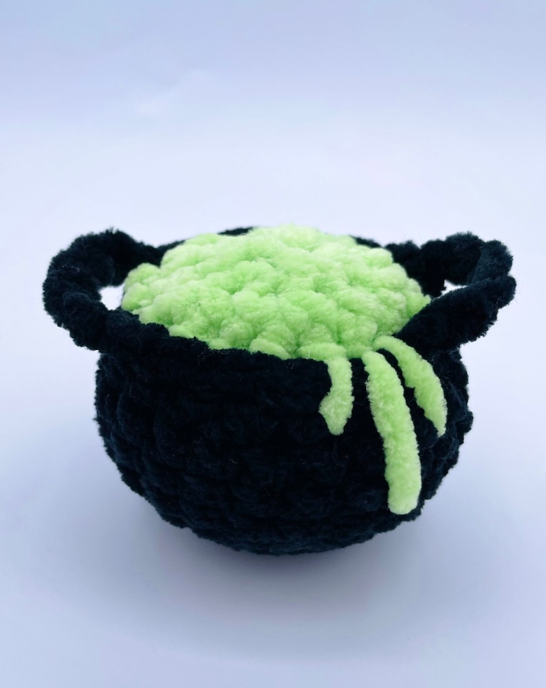 May include: A black crocheted cauldron filled with green slime. The slime is spilling over the edge of the cauldron.