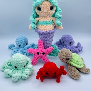 May include: A crocheted mermaid doll with a light blue tail, pink starfish, light blue, purple, and green octopus, and a red crab. The mermaid doll is wearing a light green top and has light blue eyes.