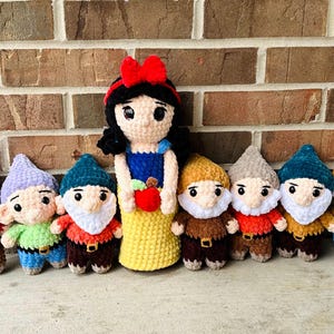May include: A group of handmade crochet dolls depicting Snow White and the seven dwarfs. Snow White is in a blue and yellow dress, holding a red apple. The dwarfs have colourful hats and outfits, with white beards.