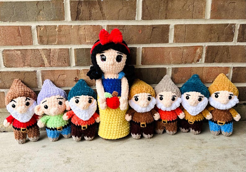 May include: A collection of handmade crochet dolls featuring Snow White and seven dwarfs. Snow White wears a blue and yellow dress and holds a red apple. The dwarfs have colorful hats and outfits, with white beards.