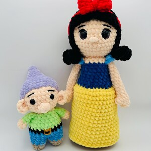 May include: Crocheted Snow White doll with a red bow and black hair, next to a crocheted dwarf doll. The Snow White doll has a blue top and yellow skirt. The dwarf doll has a green shirt, blue pants, and a purple hat.