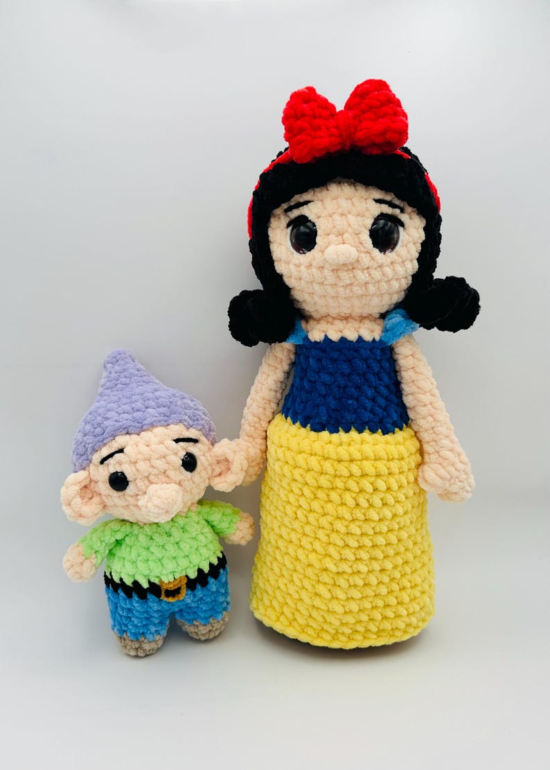 May include: Crocheted Snow White doll with a red bow and black hair, next to a crocheted dwarf doll. The Snow White doll has a blue top and yellow skirt. The dwarf doll has a green shirt, blue pants, and a purple hat.