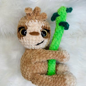 Puede incluir: Un juguete de perezoso de ganchillo hecho a mano en tonos marrones y beige, con ojos grandes y brillantes y una expresión sonriente. El perezoso está agarrando una rama verde brillante y texturizada. El peluche está sobre un fondo blanco y esponjoso.