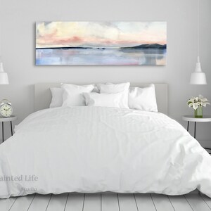 May include: A white bed with a white duvet cover and white pillows. A large abstract painting of a pink and blue sunset over a body of water hangs above the bed. The painting is framed and has a white mat.