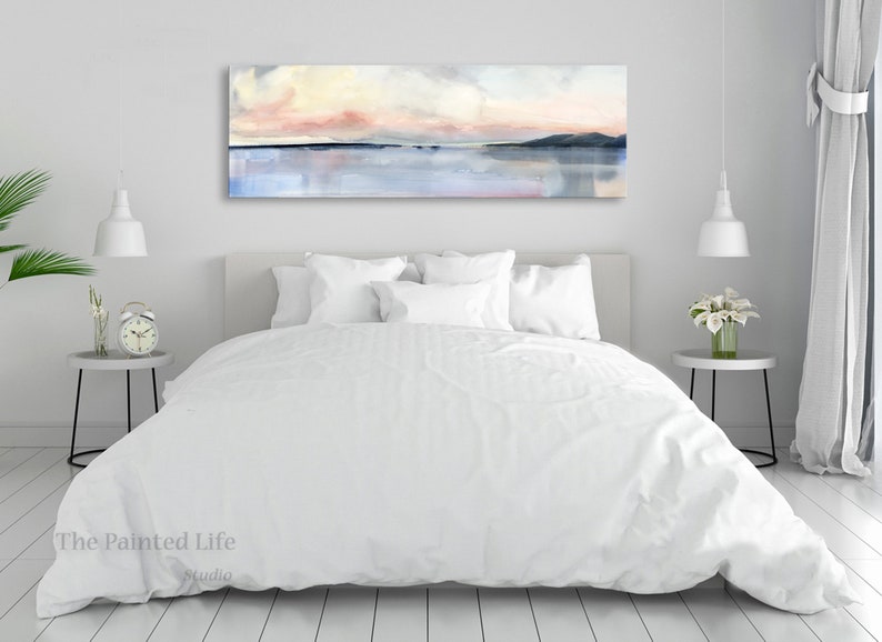 May include: A white bed with a white duvet cover and white pillows. A large abstract painting of a pink and blue sunset over a body of water hangs above the bed. The painting is framed and has a white mat.