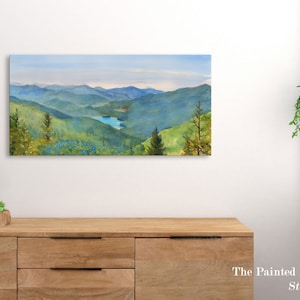 May include: A watercolor painting of a mountain range with a small lake in the distance. The mountains are a deep green and blue, and the sky is a light blue. The painting is framed and hanging on a white wall.