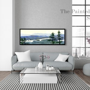 May include: A framed watercolor painting of a landscape with mountains, a river, and trees. The painting is hanging on a gray wall above a light gray sofa in a modern living room. The text "The Painted Life Studio" is visible in the upper right corner of the image.