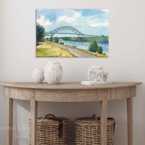 May include: Watercolor painting of a bridge over a river under a blue sky with clouds. The bridge is gray, and the landscape includes green trees and a yellow-green field. The painting is displayed above a wooden console table with decorative vases and baskets.