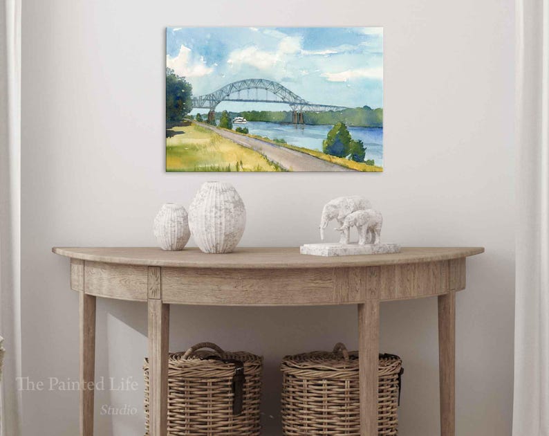 May include: Watercolor painting of a bridge over a river under a blue sky with clouds. The bridge is gray, and the landscape includes green trees and a yellow-green field. The painting is displayed above a wooden console table with decorative vases and baskets.
