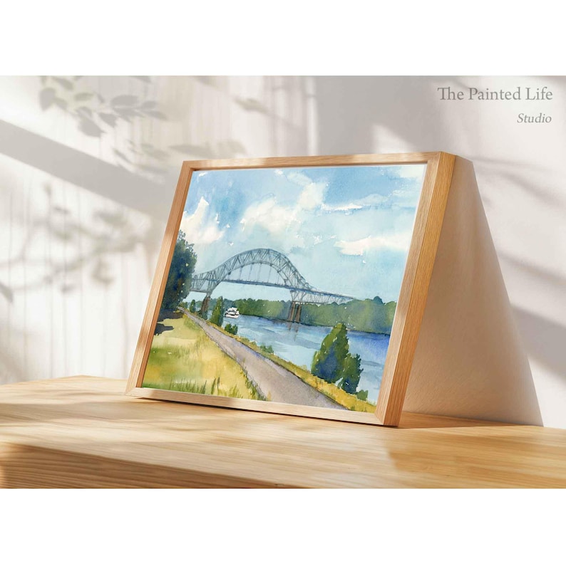 May include: Watercolor painting of a bridge over a river, framed in a light wood frame. The artwork depicts a blue sky with clouds, a green landscape, and a road. The text "The Painted Life Studio" is visible.