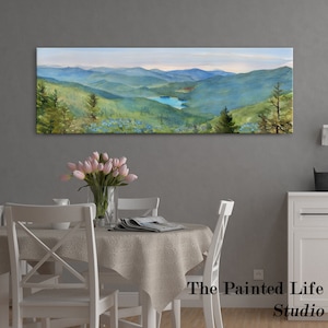 May include: A panoramic landscape painting of a mountain range with a lake in the distance. The painting is in a light blue, green, and brown color palette. The painting is hanging on a gray wall above a white table with white chairs. The text "The Painted Life Studio" is visible in the bottom right corner of the image.
