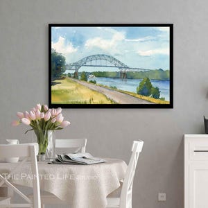 May include: Watercolor painting of a bridge over a river, framed in black. The artwork depicts a blue sky with clouds, a green landscape, and a road. The bridge is gray, and the water is blue. The painting is displayed on a gray wall.