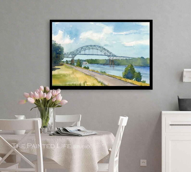 May include: Watercolor painting of a bridge over a river, framed in black. The artwork depicts a blue sky with clouds, a green landscape, and a road. The bridge is gray, and the water is blue. The painting is displayed on a gray wall.
