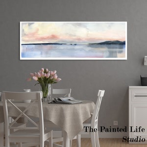 May include: A watercolor painting of a seascape with a pink and yellow sky, a blue body of water, and a dark green mountain range in the distance. The painting is framed and hanging on a gray wall above a white dining table with a white tablecloth and four white chairs. The table is decorated with a vase of pink tulips and a magazine. The text "The Painted Life Studio" is visible in the bottom right corner of the image.